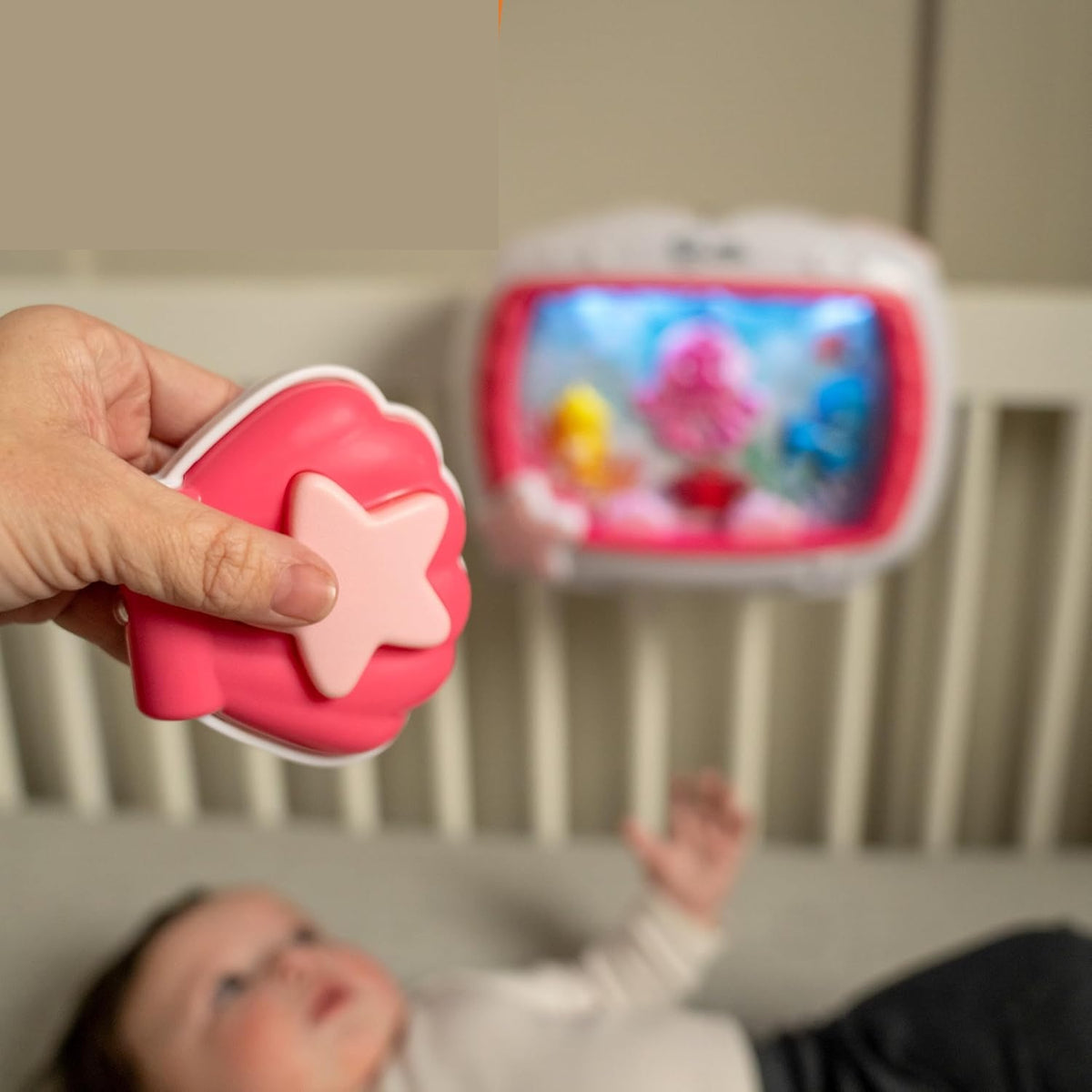 Dłoń trzymająca różowy pilot z gwiazdą, z pozytywką Baby Einstein w tle.