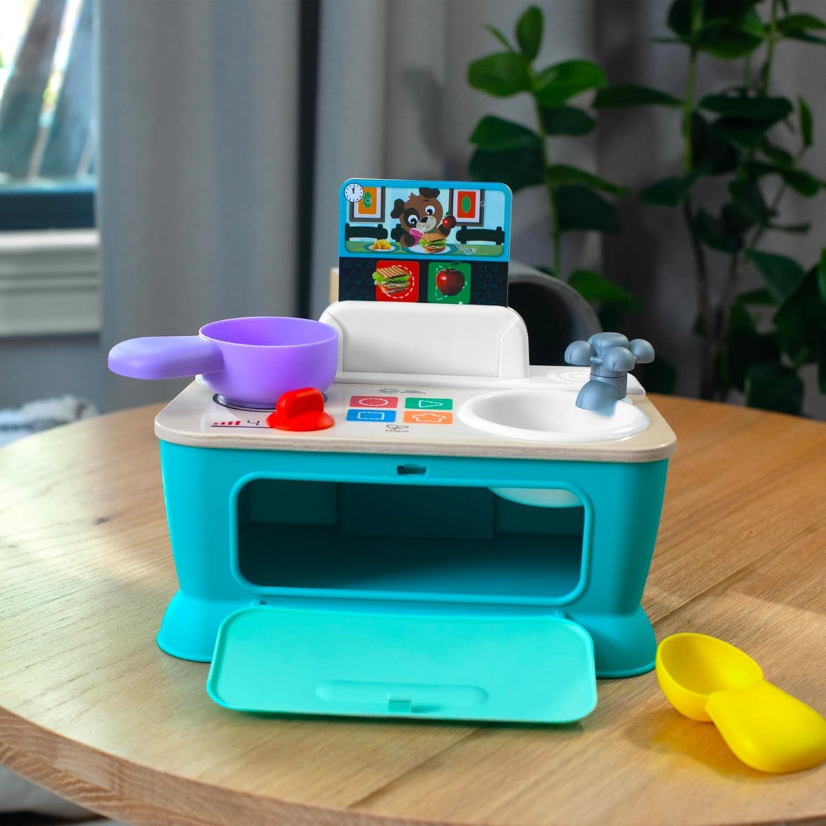 Kuchnia Baby Einstein Hape na stole z otwartymi przednimi drzwiczkami i łyżką obok.
